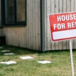 A modern wooden house with a prominent 'House for Rent' sign in the green yard.